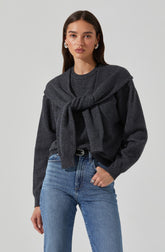 A woman wearing a dark gray sweater with a matching gray sweater tied around her shoulders. She is also wearing light blue high-waisted jeans with a black belt.